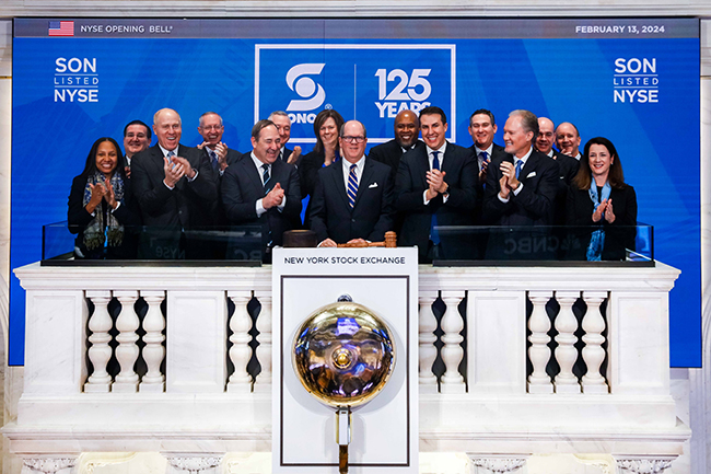Un groupe d’hommes d’affaires portant des costumes souriant et applaudissant devant un mur indiquant «&nbsp;SON LISTED NYSE, SONOCO 125 Years&nbsp;» debout devant la cloche de la Bourse de New York.
